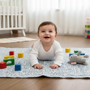 Baby lying on the Blue Rose Meadow hand block printed quilt, smiling during playtime in a bright nursery. The quilt features soft blue rose floral motifs on an ivory background with gentle hand quilting, creating a cushioned and comfortable surface. Made from breathable cotton, the quilt is ideal for baby play, tummy time, or nursery décor, combining traditional craftsmanship with softness, safety, and timeless style.