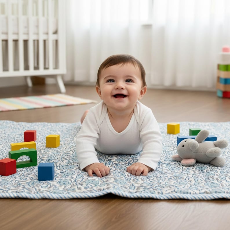 Baby lying on the Blue Rose Meadow hand block printed quilt, smiling during playtime in a bright nursery. The quilt features soft blue rose floral motifs on an ivory background with gentle hand quilting, creating a cushioned and comfortable surface. Made from breathable cotton, the quilt is ideal for baby play, tummy time, or nursery décor, combining traditional craftsmanship with softness, safety, and timeless style.