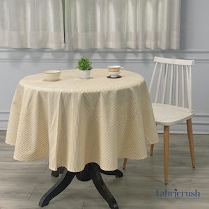 Puede incluir: Una mesa redonda cubierta con un mantel a rayas beige y blanco. Una pequeña planta en maceta, un cuenco y una taza de té con un platillo están sobre la mesa. Una silla blanca con respaldo de listones está al lado de la mesa.