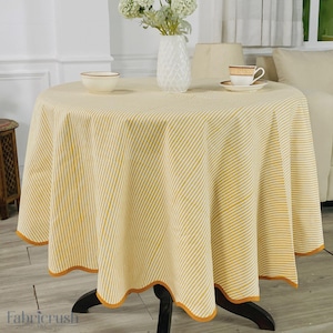 Puede incluir: Un mantel redondo con un estampado de rayas amarillas y blancas. El mantel tiene un borde festoneado con un fino borde naranja. Un cuenco, una taza y un platillo, y un jarrón con flores están sobre la mesa.