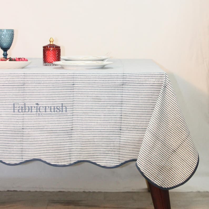 Handmade Blue striped linen tablecloth with Blue piping, styled on a dining table. The tablecloth features fine horizontal Blue and white stripes, a softly scalloped edge, and matching Blue trim for a refined finish. Displayed with white dinnerware and cutlery, it creates a clean, elegant tablescape ideal for everyday dining, farmhouse interiors, festive gatherings, and casual entertaining.