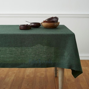 Puede incluir: Un mantel rectangular verde oscuro con una superficie texturizada. Varios cuencos de madera de diferentes tamaños están colocados sobre la mesa. La mesa tiene patas de madera de color claro y está sobre un suelo de madera.