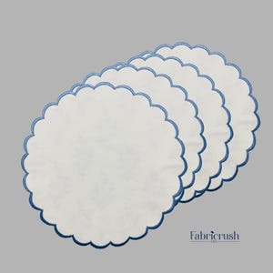 Op de afbeelding: Een stapel van vier witte placemats met geschulpte randen en een blauwe geborduurde rand. De placemats zijn diagonaal geplaatst tegen een grijze achtergrond. Het Fabricrush USA-logo staat rechtsonder.
