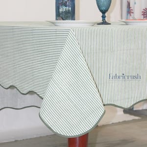May include: A green and white striped tablecloth with a scalloped edge. The tablecloth is draped over a table, with the Fabricrush logo visible in the corner. The table is set with plates and blue glassware.