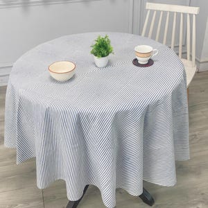 Puede incluir: Un mantel redondo con un patrón de rayas azules y blancas cubre una mesa. Una pequeña planta en maceta, un cuenco y una taza de té con un platillo están sobre la mesa. Una silla blanca está en el fondo.