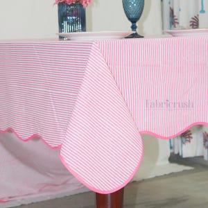 May include: A pink and white striped tablecloth with a scalloped edge and a pink trim. The tablecloth is draped over a wooden table, with tableware and floral arrangements visible in the background.
