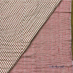 Puede incluir: Textil acolchado con dos patrones distintos. Un lado presenta un patrón repetido de pequeños motivos florales rojos sobre un fondo crema. El otro lado tiene rayas verticales rojas sobre un fondo crema. Los bordes están cuidadosamente acabados.