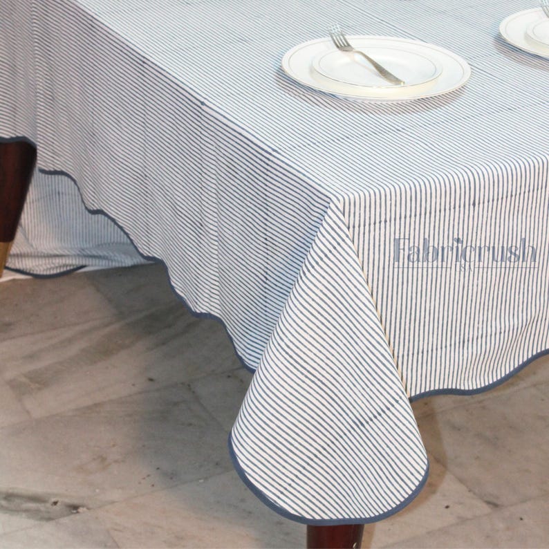 Handmade Blue striped linen tablecloth with Blue piping, draped over a dining table. The tablecloth features fine vertical Blue and white stripes with a soft scalloped edge finished in matching Blue trim. Styled with plates and glassware, it showcases a natural, elegant look ideal for everyday dining, farmhouse decor, weddings, and casual entertaining. Crafted from breathable, durable linen for a timeless tablescape.