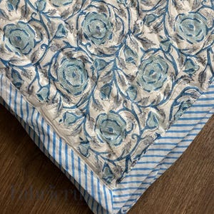 Close-up view of the Blue Rose Meadow hand block printed quilt highlighting detailed blue rose floral motifs on an ivory base. The quilt showcases traditional artisan block printing with soft blue and grey vines, fine hand quilting stitches, and a contrasting blue-and-white striped border. Made from breathable cotton, the textured surface and neat corner finish emphasize craftsmanship, comfort, and timeless design suitable for elegant bedroom decor.