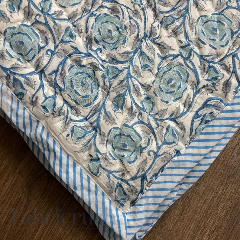Close-up view of the Blue Rose Meadow hand block printed quilt highlighting detailed blue rose floral motifs on an ivory base. The quilt showcases traditional artisan block printing with soft blue and grey vines, fine hand quilting stitches, and a contrasting blue-and-white striped border. Made from breathable cotton, the textured surface and neat corner finish emphasize craftsmanship, comfort, and timeless design suitable for elegant bedroom decor.