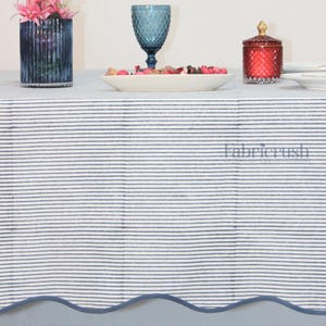 Blue striped linen tablecloth with Blue piping draped over a dining table, featuring fine Blue and white ticking stripes and a softly scalloped edge. Styled with stacked white plates, coral accent dishes, a textured blue goblet, and a floral vase, this handmade linen tablecloth adds a clean, elegant look ideal for farmhouse, coastal, and everyday dining décor.