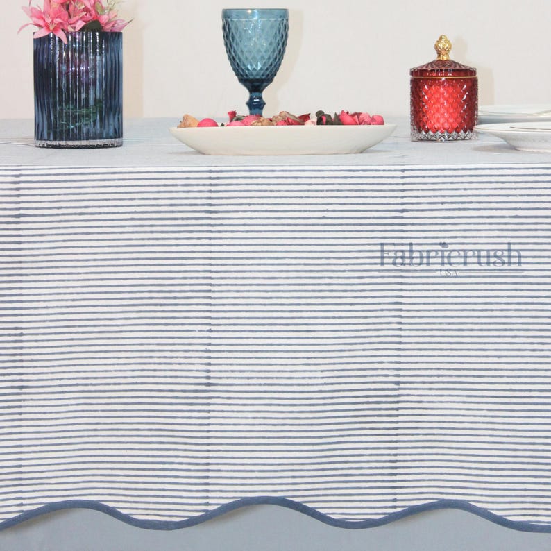 Blue striped linen tablecloth with Blue piping draped over a dining table, featuring fine Blue and white ticking stripes and a softly scalloped edge. Styled with stacked white plates, coral accent dishes, a textured blue goblet, and a floral vase, this handmade linen tablecloth adds a clean, elegant look ideal for farmhouse, coastal, and everyday dining décor.