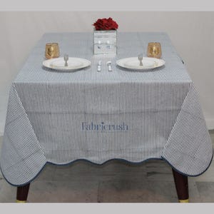 Handmade Blue striped linen tablecloth with Blue piping, displayed on a square dining table. The tablecloth features fine Blue and white ticking stripes, a softly scalloped hem, and coordinating Blue trim for a polished look. Styled with white plates, silver cutlery, and a decorative candle, it adds a fresh, timeless touch to farmhouse, coastal, and everyday dining spaces.
