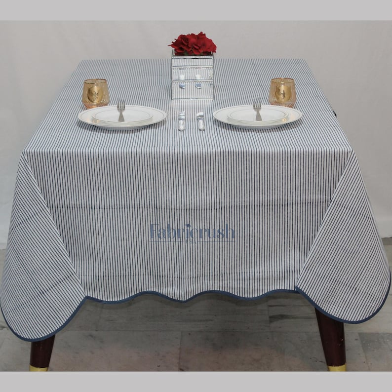 Handmade Blue striped linen tablecloth with Blue piping, displayed on a square dining table. The tablecloth features fine Blue and white ticking stripes, a softly scalloped hem, and coordinating Blue trim for a polished look. Styled with white plates, silver cutlery, and a decorative candle, it adds a fresh, timeless touch to farmhouse, coastal, and everyday dining spaces.