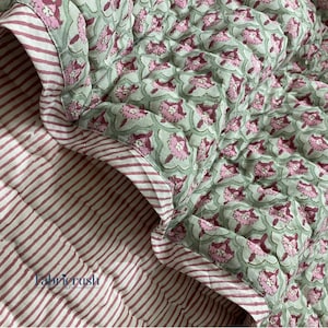 Puede incluir: Una colcha acolchada con un estampado floral en rosa y verde. El reverso presenta un estampado a rayas en rojo y blanco. La colcha tiene un borde cosido y está hecha de tela suave. El texto "Fabric Rush" es visible.