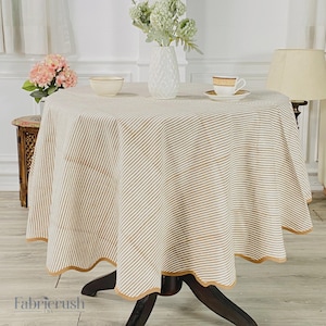 Puede incluir: Un mantel redondo con un patrón de rayas beige y blancas. El mantel está drapeado sobre una mesa de madera oscura. Un borde decorativo en un color beige más oscuro adorna el borde del mantel. Un jarrón con flores y una taza de té están sobre la mesa.