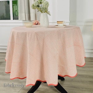 Puede incluir: Un mantel redondo con un estampado de rayas rosas y blancas. El mantel tiene un borde festoneado con un ribete coral. Un jarrón con flores blancas, un cuenco y una taza de té están sobre la mesa.
