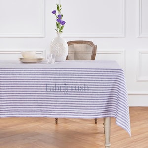 Pode incluir: Uma toalha de mesa retangular com listras horizontais em lavanda e branco. A toalha de mesa está sobre uma mesa de madeira. Um vaso branco com flores roxas, uma tigela, pratos e copos estão sobre a mesa.