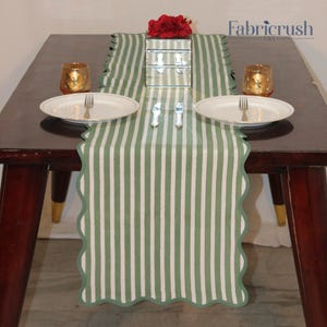 Può includere: Un set da tavolo da pranzo con un runner a righe verdi e bianche, due piatti bianchi con bordi dorati, due portacandele dorati e una scatola a specchio. Il runner ha un bordo smerlato. Il logo Fabricrush USA è visibile.