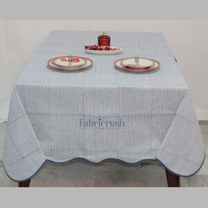 Handmade Blue striped linen tablecloth with Blue piping, displayed on a square dining table. The tablecloth features fine Blue and white ticking stripes, a softly scalloped hem, and coordinating Blue trim for a polished look. Styled with white plates, silver cutlery, and a decorative candle, it adds a fresh, timeless touch to farmhouse, coastal, and everyday dining spaces.