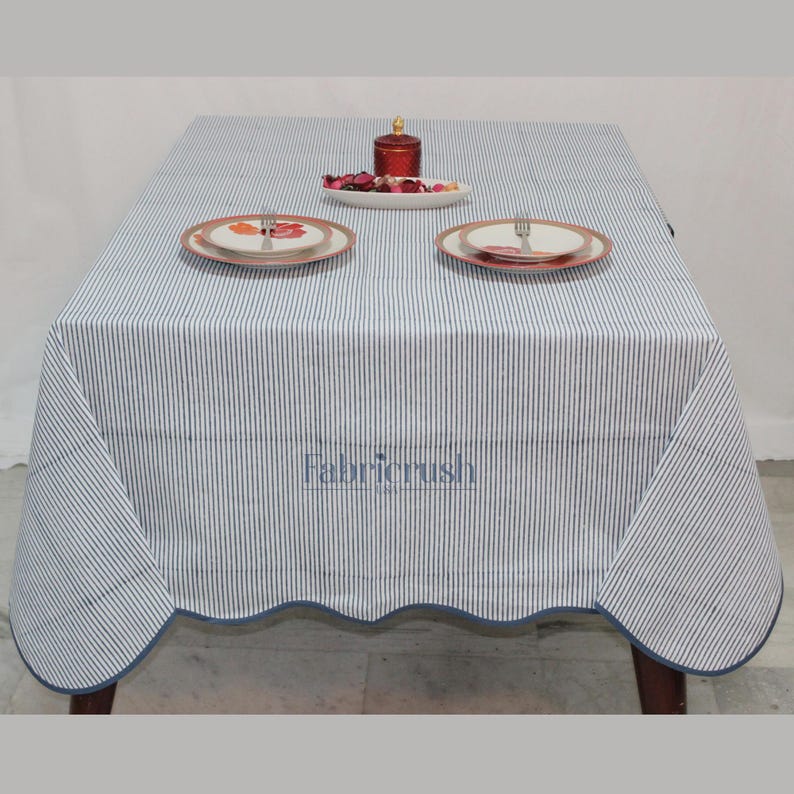 Handmade Blue striped linen tablecloth with Blue piping, displayed on a square dining table. The tablecloth features fine Blue and white ticking stripes, a softly scalloped hem, and coordinating Blue trim for a polished look. Styled with white plates, silver cutlery, and a decorative candle, it adds a fresh, timeless touch to farmhouse, coastal, and everyday dining spaces.