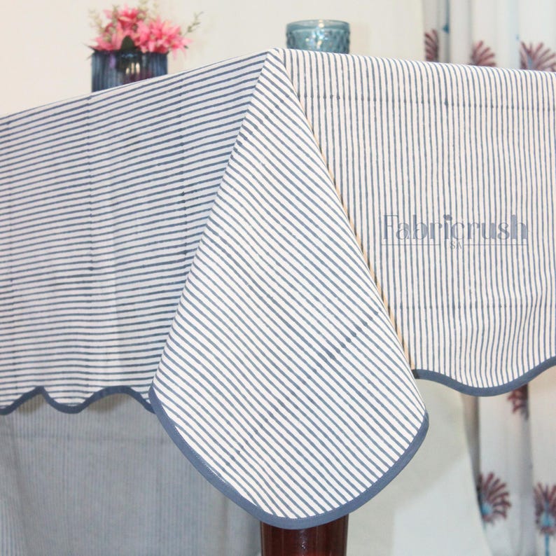 Handmade Blue striped linen tablecloth with Blue piping, draped over a dining table. The tablecloth features fine vertical Blue and white stripes with a soft scalloped edge finished in matching Blue trim. Styled with plates and glassware, it showcases a natural, elegant look ideal for everyday dining, farmhouse decor, weddings, and casual entertaining. Crafted from breathable, durable linen for a timeless tablescape.