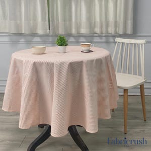 Puede incluir: Un mantel redondo con un estampado de rayas rosas y blancas cubre una mesa pequeña. Una silla blanca está a la derecha. Una pequeña planta, un cuenco y una taza de té están sobre la mesa. El mantel es de tela.