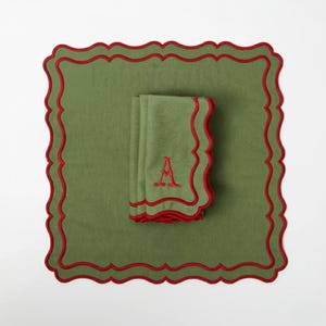 Puede incluir: Un juego de servilletas de tela verdes y un mantel individual con bordes festoneados rojos. Las servilletas están dobladas y bordadas con la letra "A" en rojo. El conjunto tiene un tema festivo.