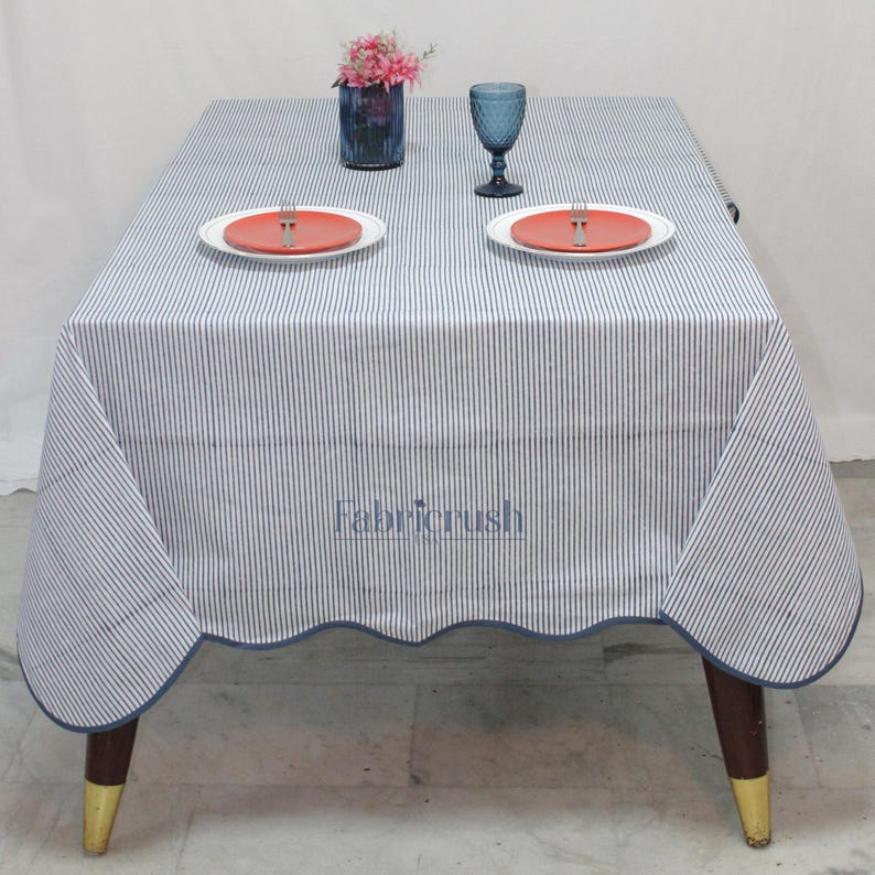 Handmade Blue striped linen tablecloth with Blue piping, displayed on a square dining table. The tablecloth features fine Blue and white ticking stripes, a softly scalloped hem, and coordinating Blue trim for a polished look. Styled with white plates, silver cutlery, and a decorative candle, it adds a fresh, timeless touch to farmhouse, coastal, and everyday dining spaces.