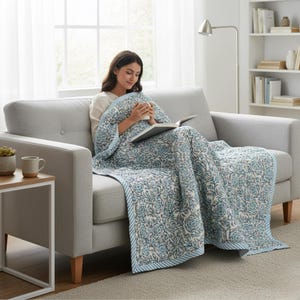 Blue Rose Meadow Hand Block Printed Quilt styled on a modern sofa, draped over a woman relaxing with a book and warm drink. The quilt features intricate blue rose and floral vine motifs on a soft ivory cotton base, finished with a blue and white striped border. The image highlights the quilt’s lightweight comfort, hand-quilted texture, reversible design, and elegant handcrafted appeal for cozy living spaces.