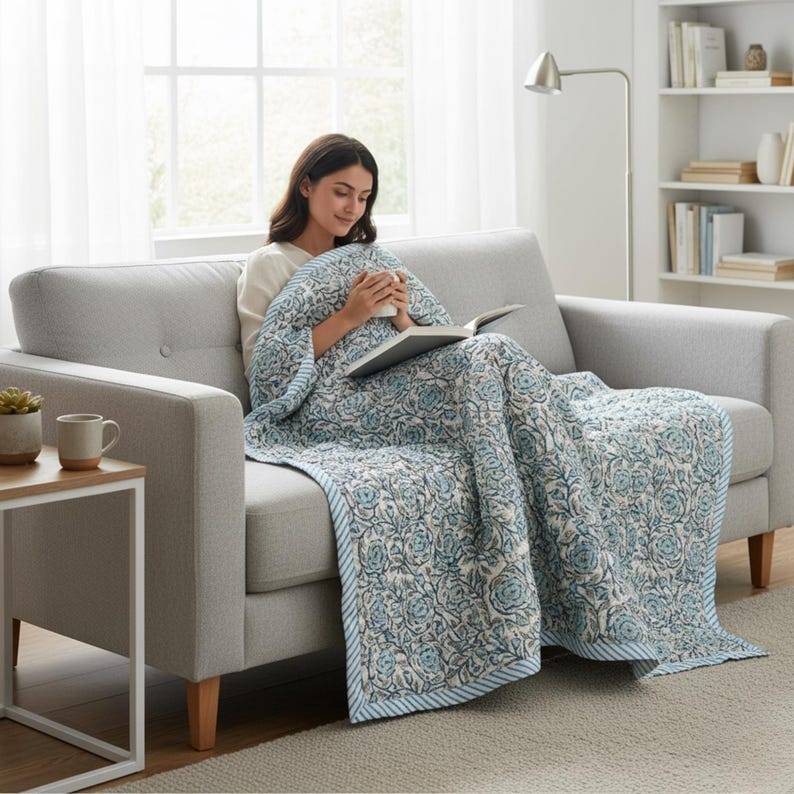 Blue Rose Meadow Hand Block Printed Quilt styled on a modern sofa, draped over a woman relaxing with a book and warm drink. The quilt features intricate blue rose and floral vine motifs on a soft ivory cotton base, finished with a blue and white striped border. The image highlights the quilt’s lightweight comfort, hand-quilted texture, reversible design, and elegant handcrafted appeal for cozy living spaces.