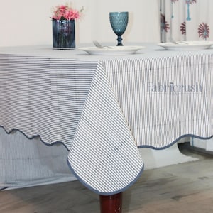 Handmade Blue striped linen tablecloth with Blue piping, draped over a dining table. The tablecloth features fine vertical Blue and white stripes with a soft scalloped edge finished in matching Blue trim. Styled with plates and glassware, it showcases a natural, elegant look ideal for everyday dining, farmhouse decor, weddings, and casual entertaining. Crafted from breathable, durable linen for a timeless tablescape.
