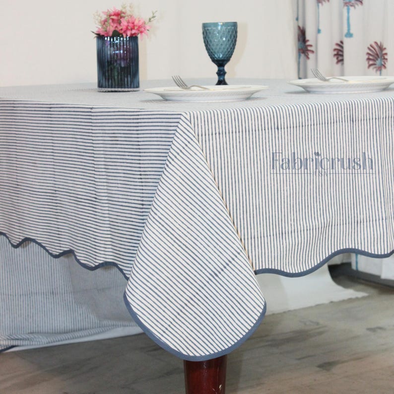 Handmade Blue striped linen tablecloth with Blue piping, draped over a dining table. The tablecloth features fine vertical Blue and white stripes with a soft scalloped edge finished in matching Blue trim. Styled with plates and glassware, it showcases a natural, elegant look ideal for everyday dining, farmhouse decor, weddings, and casual entertaining. Crafted from breathable, durable linen for a timeless tablescape.