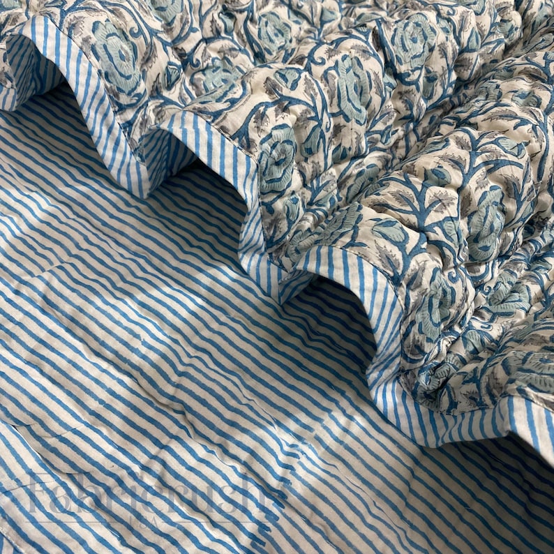 Close-up view of the Blue Rose Meadow Hand Block Printed Quilt highlighting its reversible design. The top features intricate hand-block printed blue floral vine and rose motifs on a soft ivory base, while the reverse shows delicate blue and white vertical stripes. The image emphasizes fine quilting, soft cotton texture, neatly finished edges, and handcrafted artisanal detailing for elegant, cozy bedding decor.