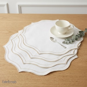 Puede incluir: Un conjunto de manteles individuales blancos con bordes festoneados y bordados en beige. Una taza y un platillo blancos, una cuchara plateada y una ramita de vegetación descansan encima. Los manteles individuales están sobre una superficie de madera clara.