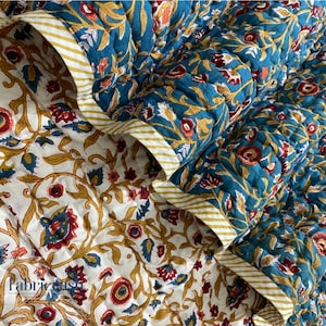 Op de afbeelding: Gewatteerde stof met een bloemenpatroon in teal, rood en goud op een witte achtergrond. De randen zijn afgewerkt met een gestreepte goud-witte rand. De stof is gevouwen en toont beide zijden van het ontwerp.