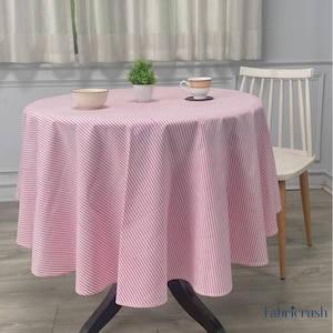 Puede incluir: Un mantel redondo a rayas rosas y blancas cubre una mesa de madera oscura. Una pequeña planta, un cuenco y una taza de té están sobre la mesa. Una silla blanca está en el fondo.