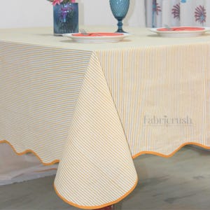 以下が含まれることがあります： 黄と白のストライプ柄の長方形のテーブルクロス。 テーブルクロスは、オレンジ色のトリムが付いたスカラップエッジです。 テーブルには、お皿、カトラリー、青いグラスがセットされています。