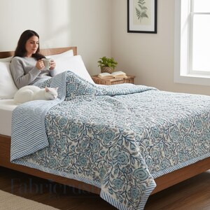 Blue Rose Meadow Hand Block Printed Quilt styled on a cozy bed, featuring intricate blue floral rose and vine motifs on a soft ivory base. The quilt is finished with a blue and white striped border and matching reverse, shown in a serene bedroom setting with a woman relaxing with a warm drink and a sleeping white cat, highlighting comfort, handcrafted detailing, and elegant, nature-inspired bedding décor.