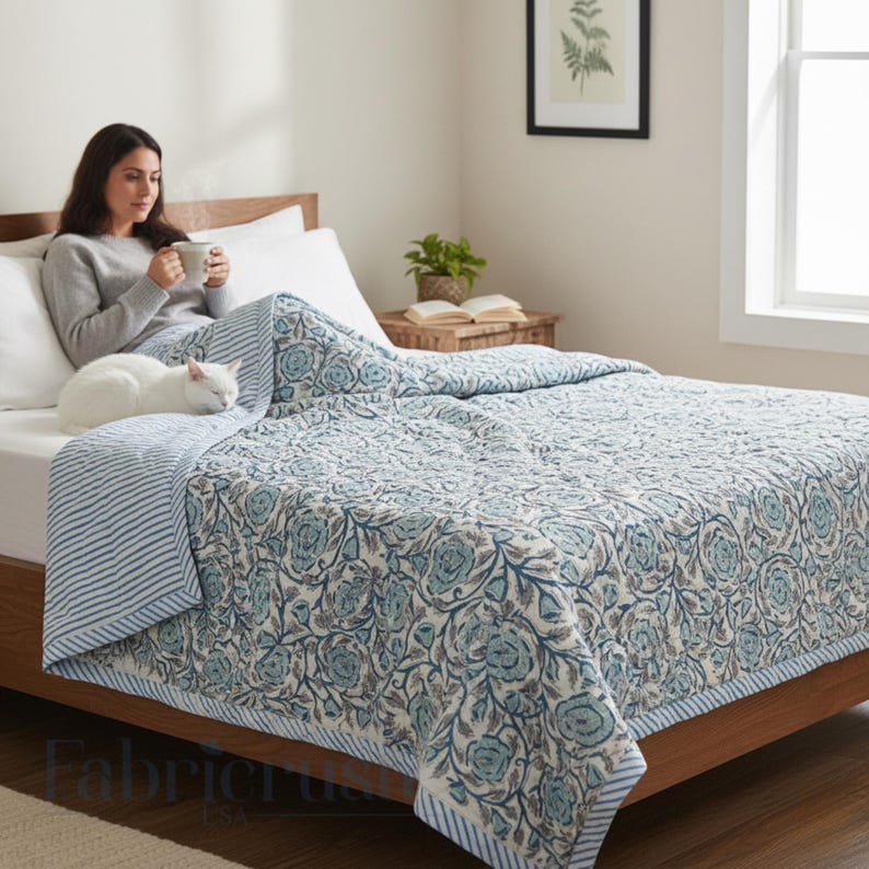 Blue Rose Meadow Hand Block Printed Quilt styled on a cozy bed, featuring intricate blue floral rose and vine motifs on a soft ivory base. The quilt is finished with a blue and white striped border and matching reverse, shown in a serene bedroom setting with a woman relaxing with a warm drink and a sleeping white cat, highlighting comfort, handcrafted detailing, and elegant, nature-inspired bedding décor.