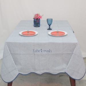 Handmade Blue striped linen tablecloth with Blue piping, displayed on a square dining table. The tablecloth features fine Blue and white ticking stripes, a softly scalloped hem, and coordinating Blue trim for a polished look. Styled with white plates, silver cutlery, and a decorative candle, it adds a fresh, timeless touch to farmhouse, coastal, and everyday dining spaces.