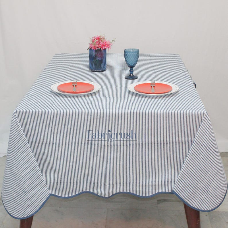 Handmade Blue striped linen tablecloth with Blue piping, displayed on a square dining table. The tablecloth features fine Blue and white ticking stripes, a softly scalloped hem, and coordinating Blue trim for a polished look. Styled with white plates, silver cutlery, and a decorative candle, it adds a fresh, timeless touch to farmhouse, coastal, and everyday dining spaces.