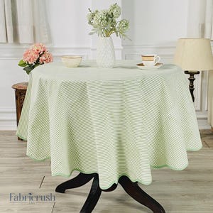 Puede incluir: Un mantel redondo con un diseño de rayas verdes y blancas. El mantel está drapeado sobre una mesa de madera oscura. El borde del mantel tiene un diseño festoneado con un ribete verde. Un jarrón de flores y una lámpara están en el fondo.