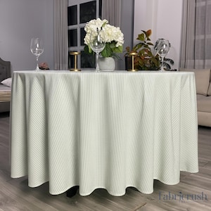 Può includere: Una tovaglia rotonda a righe bianche e verde chiaro copre un tavolo. Il tavolo è apparecchiato con calici, fiori e candele decorative. La tovaglia ha un bordo smerlato ed è realizzata in un tessuto morbido.