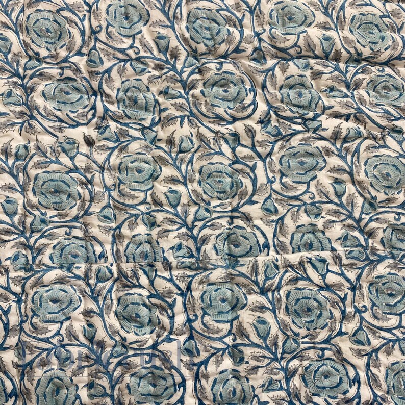Close-up detail of the Blue Rose Meadow Hand Block Printed Quilt featuring repeating blue rose blooms and leafy vine motifs printed on a soft ivory cotton fabric. The image highlights hand-quilted stitching, gentle texture, and artisanal block print variations, emphasizing the quilt’s handcrafted quality, elegant floral pattern, and timeless, nature-inspired bedding design.