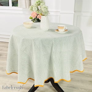 Puede incluir: Un mantel redondo con un patrón de rayas verdes y blancas. El mantel tiene un borde festoneado con un ribete amarillo. Un jarrón con flores, una taza de té y un cuenco están sobre la mesa. El logotipo de Fabricrush USA es visible.