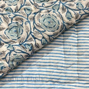 Close-up view of the Blue Rose Meadow Hand Block Printed Quilt showing its reversible design. One side features detailed blue rose and floral vine motifs hand block printed on a soft ivory cotton base, while the reverse displays delicate blue and white striped patterns. The image highlights fine hand quilting, soft texture, artisanal craftsmanship, and elegant, timeless bedding appeal.