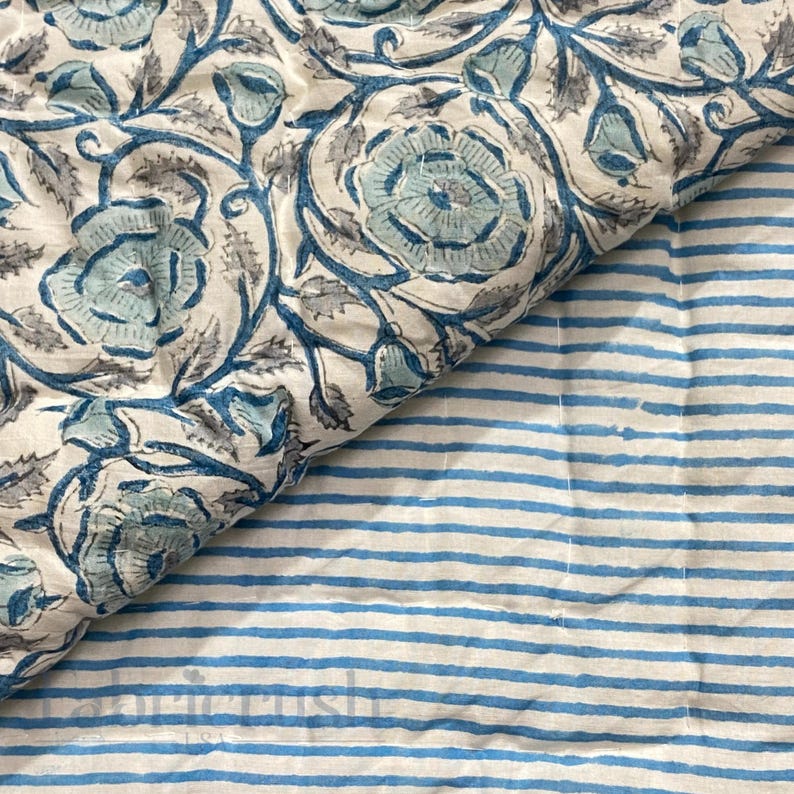 Close-up view of the Blue Rose Meadow Hand Block Printed Quilt showing its reversible design. One side features detailed blue rose and floral vine motifs hand block printed on a soft ivory cotton base, while the reverse displays delicate blue and white striped patterns. The image highlights fine hand quilting, soft texture, artisanal craftsmanship, and elegant, timeless bedding appeal.