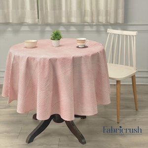 Puede incluir: Una mesa redonda cubierta con un mantel a rayas rojas y blancas. Una pequeña planta en maceta, un cuenco y una taza de té con un platillo están sobre la mesa. Una silla blanca con patas de madera está al lado de la mesa.