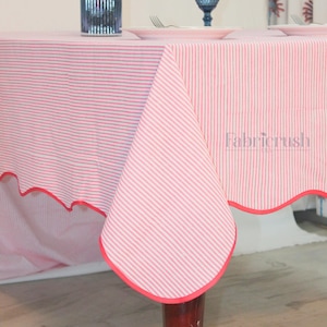 Op de afbeelding: Een roze en witte gestreepte tafelkleed met een geschulpte rand in rood. Het tafelkleed is over een tafel gedrapeerd, met een glimp van een tafelsetting met borden en bestek.